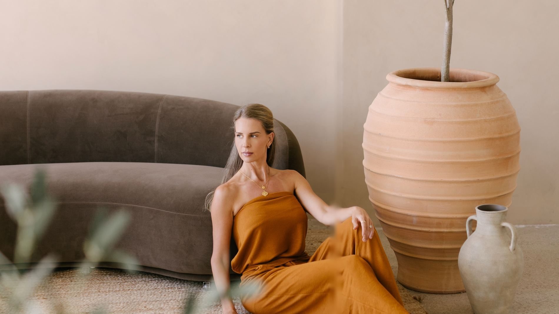 Elle Wilk seated on the floor beside an olive tree and terracotta urn in a warm, sunlit room