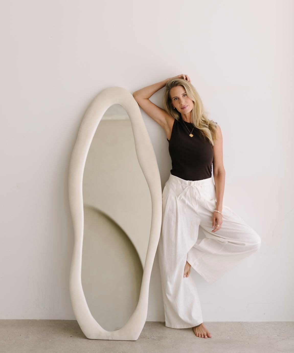 Elle leaning against the wall beside a tall sculptural mirror